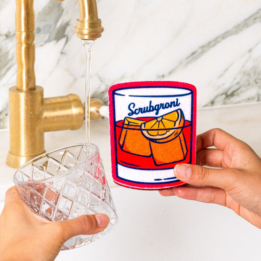 person holding scrubgroni negroni kitchen sponge washing rocks glass in kitchen sink under running water