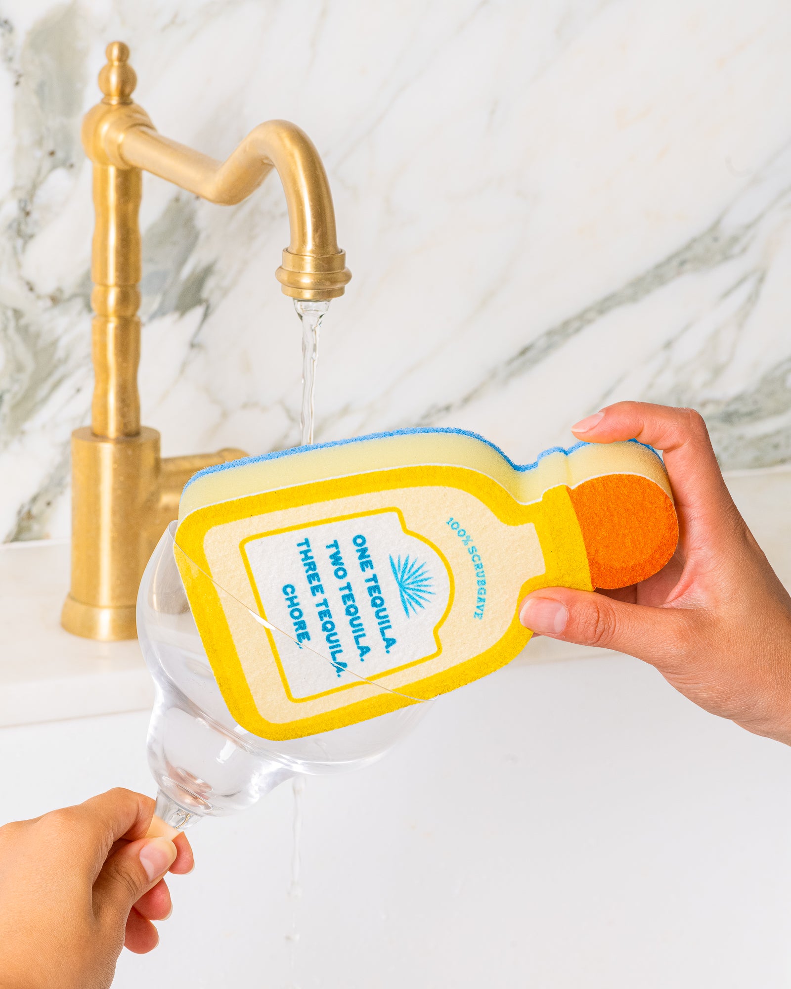 Hand holding a sponge shaped like a bottle of tequila next to a glass under a running faucet.