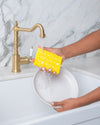 yellow kitchen sponge washing dishes in sink. sponge has a wine bottle print. biodegradable compressed kitchen sponge.