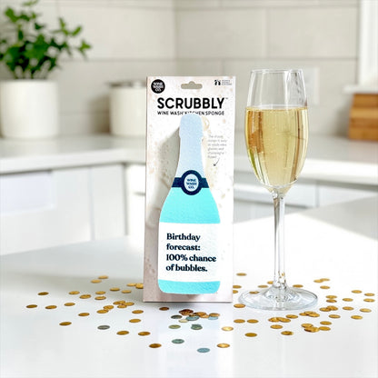 Scrubbly wine bottle-shaped kitchen sponge with a glass of champagne and confetti on a kitchen counter.
